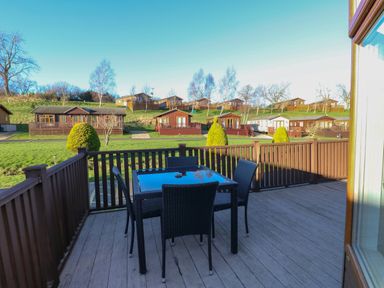 An outdoor deck with a table and four chairs overlooking multiple cabins on a grassy hillside at The Sanctuary Badgers Retreat Holiday Park near Catterick Garrison North Yorkshire