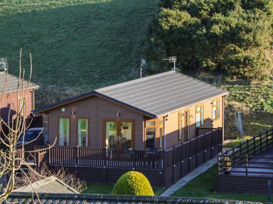 A wooden cabin with a fenced porch and green hillside in the background at The Sanctuary in Badgers Retreat Holiday Park near Catterick Garrison North Yorkshire