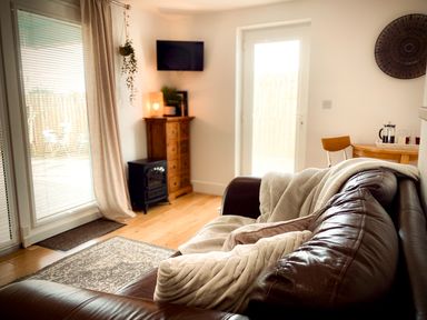 A living room with a leather sofa with cushions and blankets a wooden cabinet a wall-mounted TV and a glass door at The Studio in Crackington Haven