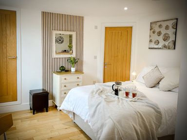 A bedroom with a bed and a tray on it a chest of drawers with a plant and mirror above and two wooden doors at The Studio in Crackington Haven
