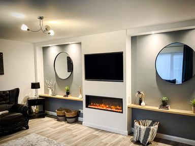 A living room with a black leather armchair shelves with decorative items a TV above a fireplace and two round mirrors at Trelawne Near Crackington Haven