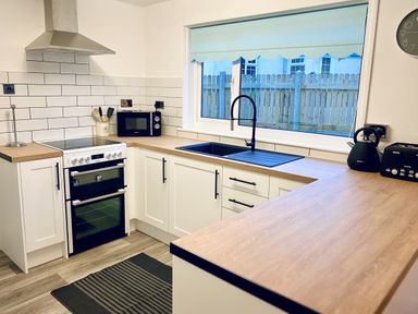 A kitchen with white cabinets wooden countertops a black sink a stove a microwave a kettle and a toaster at Trelawne Near Crackington Haven