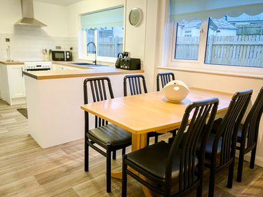 A dining area with a wooden table and six black chairs next to a kitchen with white cabinets and appliances at Trelawne Near Crackington Haven