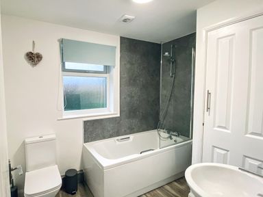 A bathroom with a bathtub shower, toilet, window with blind, sink, and a door at Trelawne Near Crackington Haven