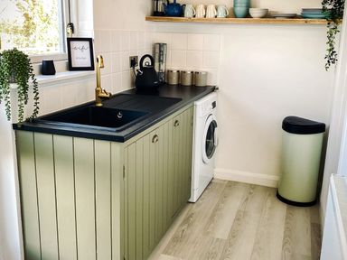 A kitchen area with a black sink gold faucet green cabinets washing machine and shelving with dishes at Penkenna in Crackington Haven