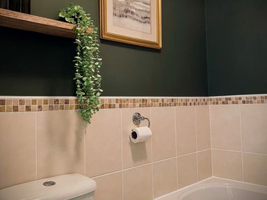 A bathroom with beige tiled walls a toilet a toilet paper holder a framed picture and a green plant at Penkenna in Crackington Haven