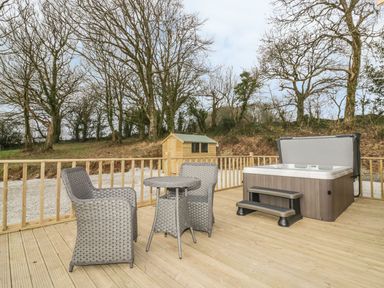 An outdoor deck with a hot tub a small table and two chairs and a wooden shed surrounded by leafless trees