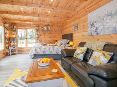 A wooden cabin interior with a bed a black leather sofa a wooden coffee table and large windows at Little Orchard Lodge in Goonhavern