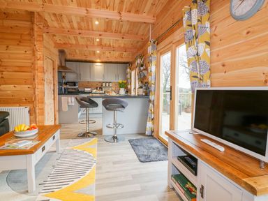 A wooden cabin interior with a kitchen island and bar stools a coffee table with fruit a television on a stand and glass doors with curtains at Little Orchard Lodge in Goonhavern