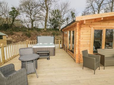An outdoor wooden deck with wicker furniture and a hot tub attached to a wooden cabin at Little Orchard Lodge in Goonhavern