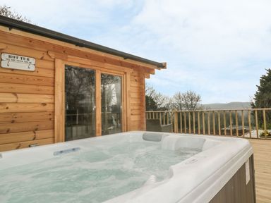 An outdoor hot tub on a wooden deck next to a wooden cabin with glass doors at Little Orchard Lodge in Goonhavern