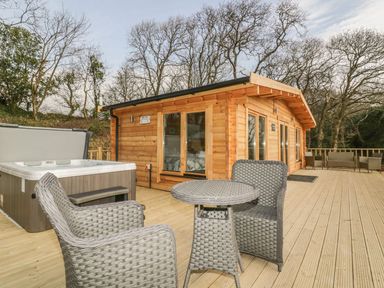 A wooden cabin with sliding glass doors on a large deck with wicker chairs a table and a hot tub at Little Orchard Lodge in Goonhavern