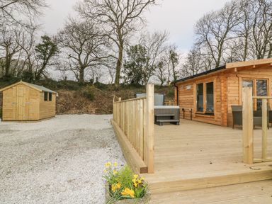 A gravel area with a wooden shed and a wooden deck attached to a cabin with garden chairs and a hot tub at Little Orchard Lodge in Goonhavern