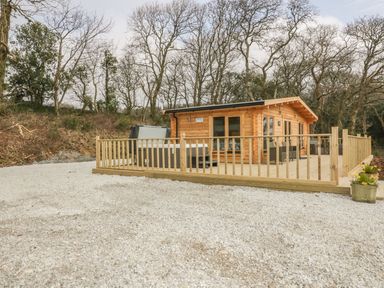 A wooden cabin with a deck and hot tub surrounded by a gravel area and trees at Little Orchard Lodge in Goonhavern