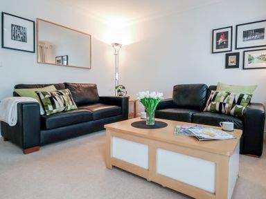 A living room with two black leather sofas a wooden coffee table with magazines and flowers and framed pictures on the wall at Bay Tree Cottage in Peasenhall