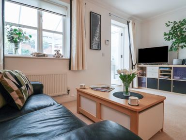 A living room with a black leather sofa a wooden coffee table with flowers a window with curtains and a television on a stand at Bay Tree Cottage in Peasenhall