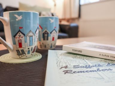 A close up of two mugs with beach hut designs and a book on a table in a living room at Bay Tree Cottage Peasenhall