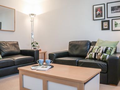 A living room with two black sofas a wooden coffee table with books and mugs a floor lamp side table with a plant and framed pictures on the wall at Bay Tree Cottage in Peasenhall
