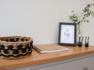 A wooden countertop with a woven basket notebook framed wifi sign and a vase with flowers at Bay Tree Cottage in Peasenhall