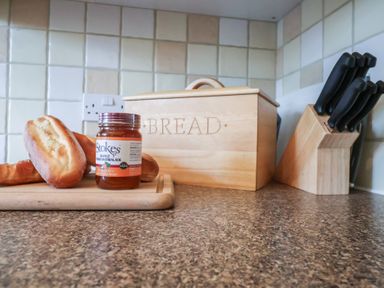 A kitchen countertop with a loaf of bread a jar of marmalade a wooden bread box and a knife block with knives at Bay Tree Cottage in Peasenhall