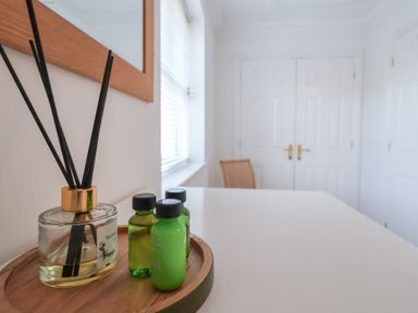 A white table with a wooden tray holding a reed diffuser and three small green bottles in a room with white walls and double white doors at Bay Tree Cottage in Peasenhall