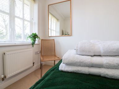 A bedroom with stacked white towels on a green bedspread next to a wicker chair and window at Bay Tree Cottage in Peasenhall
