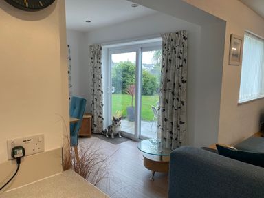 A living area with a dog sitting near glass sliding doors looking out to a garden with curtains and a blue chair at Maceley in Kingsbridge