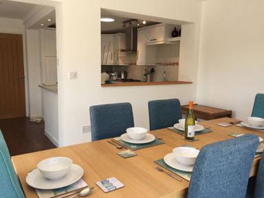 A dining area with a wooden table set with white dishes and blue chairs next to a kitchen with white cabinets at Maceley in Kingsbridge