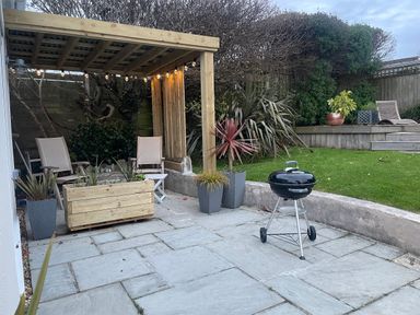 A patio with chairs under a wooden pergola a barbecue on a stone floor and plants near a grass lawn at Maceley in Kingsbridge