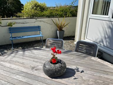 An outdoor patio with a round wooden table and chairs a flower pot with red flowers on the table a blue bench against a wall and a potted plant near a door at Maceley in Kingsbridge