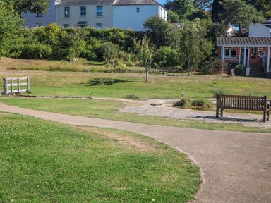 A park area with benches a small circular pond paved walkway some trees and bushes and houses in the background at Maceley in Kingsbridge