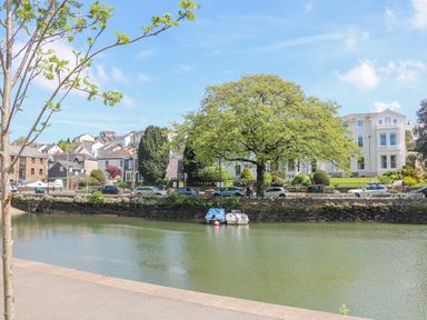 A river with boats near a stone wall and cars parked along a road with houses and a large tree behind at Maceley in Kingsbridge