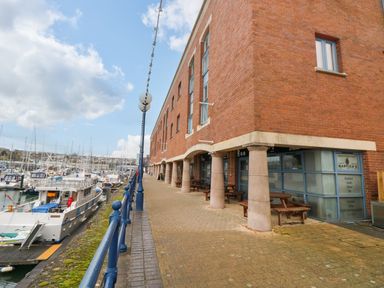 A walkway along a marina with boats docked next to a brick building with columns and outdoor seating at 15 Sovereign House in Milford Haven