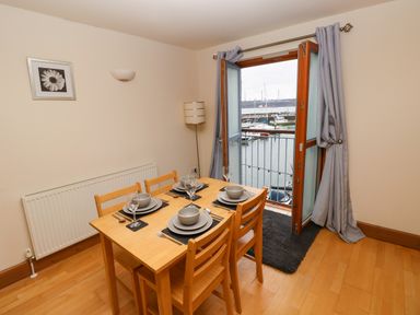 A dining area with a wooden table set for four near an open door with a view of boats on water at 15 Sovereign House in Milford Haven