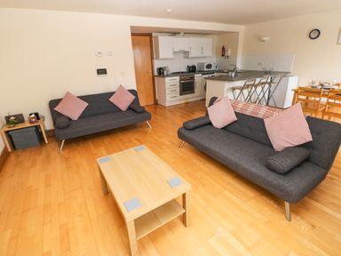 A living room with two sofas pink cushions a wooden coffee table and an open kitchen with bar stools and a dining table at 15 Sovereign House in Milford Haven