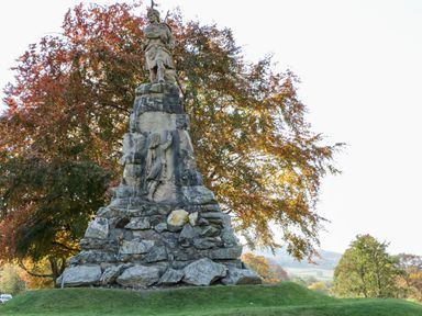 A stone statue monument with a figure on top and a relief sculpture below surrounded by trees with autumn leaves at Dundarach in Aberfeldy