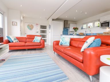 A living room with two orange sofas and blue striped rug connected to a kitchen with white cabinets and blue backsplash at Petroc in Portreath