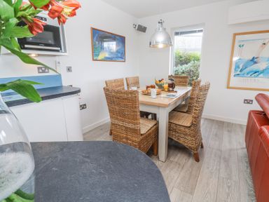 A dining area with a wooden table and six wicker chairs next to a kitchen counter and window at Petroc in Portreath