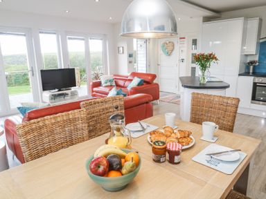 A kitchen and dining area with a wooden table set with fruit bowl pastries jars of jam and mugs and red sofas in the living room at Petroc in Portreath