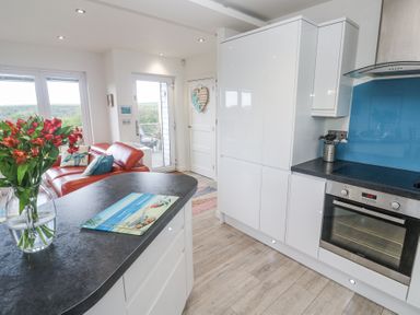 A kitchen with white cabinets and black countertop and a red sofa with cushions near a glass door and window at Petroc in Portreath