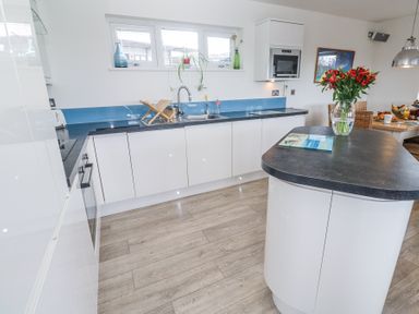 A kitchen with white cabinets black countertops a vase of red flowers on an island and windows above the sink at Petroc in Portreath