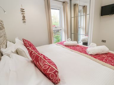A bedroom with a bed with red and beige accents rolled white towels on bed and a window and mirror at Petroc in Portreath