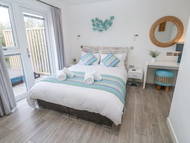 A bedroom with a double bed with striped pillows and rolled towels next to glass doors and a dressing table with a round mirror at Petroc in Portreath
