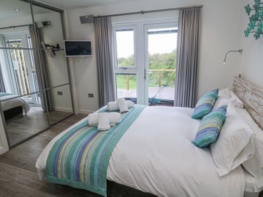 A bedroom with a double bed with striped bedding a mounted tv mirrored closet doors and glass doors leading outside at Petroc in Portreath
