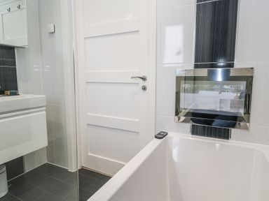 A bathroom with a bathtub a remote control a wall-mounted sink a white door and tiled walls at Petroc in Portreath