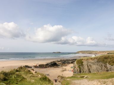 A beach with sand rocks and grass with people on the shore and an island with a lighthouse in the sea at Petroc in Portreath