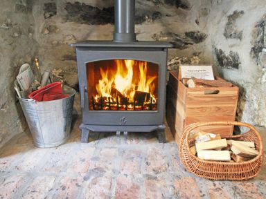 A burning wood stove with a metal bucket holding fire tools on the left and wooden logs in a basket and box on the right at Ivy House in Beddgelert