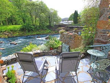 A riverside patio area with two reclining chairs and a small round table with chairs at Ivy House in Beddgelert