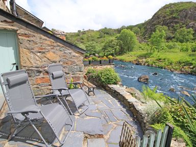 A stone patio with two reclining chairs and plants by a river at Ivy House in Beddgelert