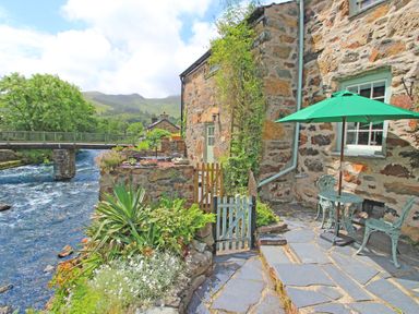 A patio with green umbrella and chairs next to a stone house by a river with a bridge in the background at Ivy House in Beddgelert